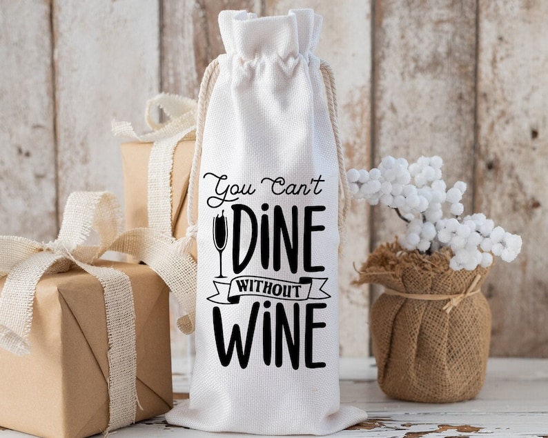 May include: White cotton wine bag with black text that reads "You Can't Dine Without Wine". The bag has a drawstring closure and is sitting on a wooden surface.