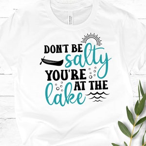 May include: White t-shirt with black text that reads "Don't be salty you're at the lake" with a turquoise blue graphic of a canoe, sun, and waves.