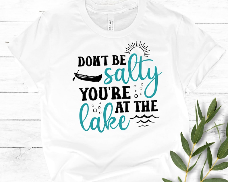 May include: White t-shirt with black text that reads "Don't be salty you're at the lake" with a turquoise blue graphic of a canoe, sun, and waves.