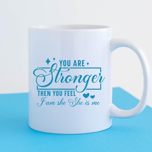May include: White ceramic mug with a blue inscription that reads "You are stronger than you feel I am she she is me". The inscription is framed by a rectangle and two small hearts.