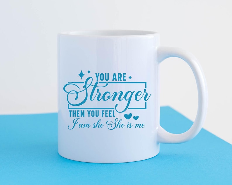 May include: White ceramic mug with a blue inscription that reads "You are stronger than you feel I am she she is me". The inscription is framed by a rectangle and two small hearts.