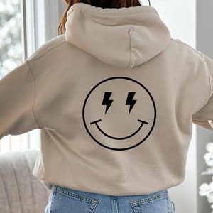 K&ouml;nnte beinhalten: Ein beiger Hoodie mit einem schwarzen Smiley-Motiv mit Blitzen als Augen. Der Hoodie hat eine Kapuze mit Kordelzug.