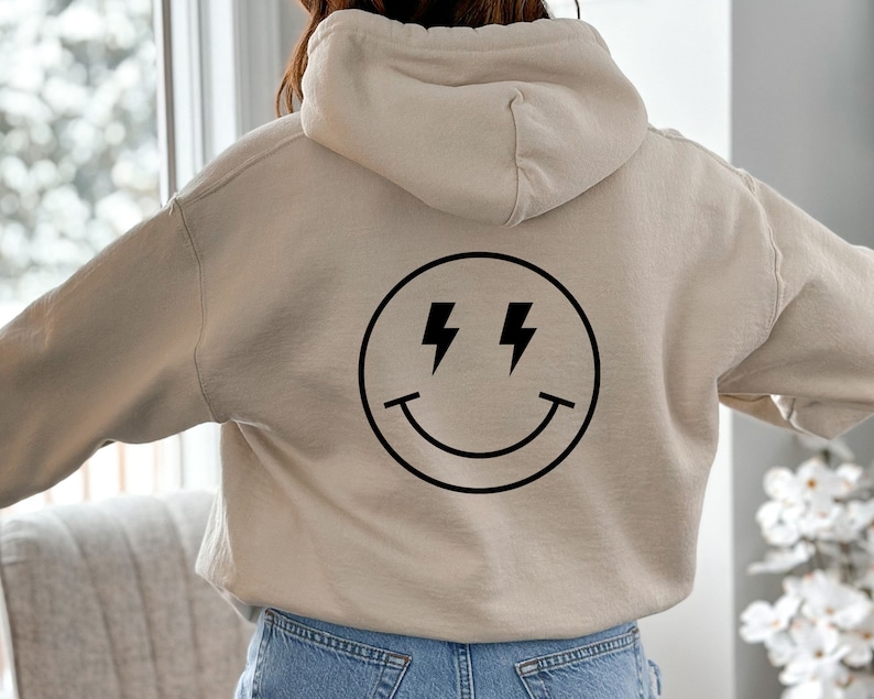 K&ouml;nnte beinhalten: Ein beiger Hoodie mit einem schwarzen Smiley-Motiv mit Blitzen als Augen. Der Hoodie hat eine Kapuze mit Kordelzug.