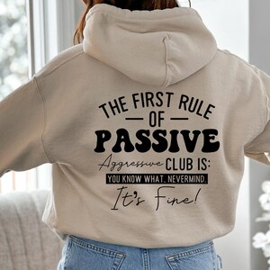 May include: A beige hoodie with a black graphic print that reads "The First Rule - Of - Passive Aggressive Club Is: You Know What, Nevermind. It's Fine!"