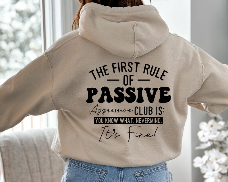 May include: A beige hoodie with a black graphic print that reads "The First Rule - Of - Passive Aggressive Club Is: You Know What, Nevermind. It's Fine!"