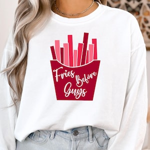 Op de afbeelding: Wit langarm shirt met een rode en roze afbeelding van frietjes in een rode bak. De tekst "Fries Before Guys" is in wit op de bak gedrukt.