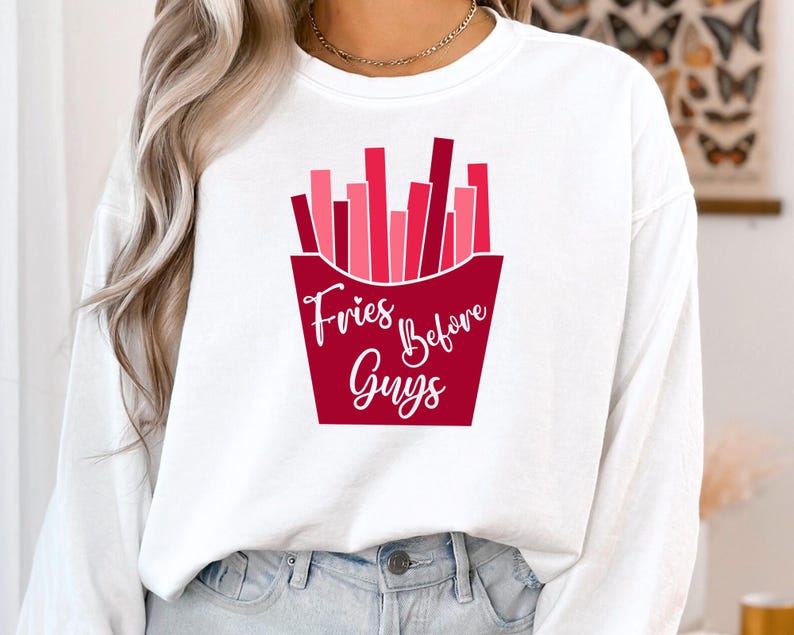 Op de afbeelding: Wit langarm shirt met een rode en roze afbeelding van frietjes in een rode bak. De tekst "Fries Before Guys" is in wit op de bak gedrukt.