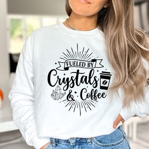 May include: White sweatshirt with black text that reads "Fueled by Crystals & Coffee". The text is surrounded by a sunburst design and a coffee cup graphic.