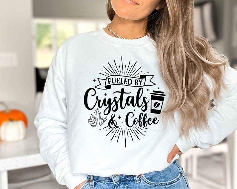 May include: White sweatshirt with black text that reads "Fueled by Crystals & Coffee". The text is surrounded by a sunburst design and a coffee cup graphic.