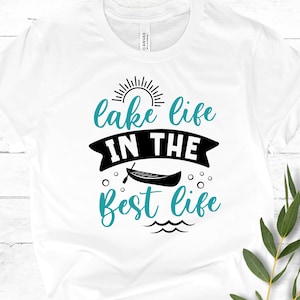 May include: White t-shirt with black text that reads "lake life in the best life". The text is surrounded by a sun, a canoe, and water waves.