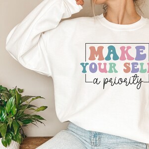 May include: White sweatshirt with the text "MAKE YOUR SELF - a priority" in colorful, retro-style lettering.