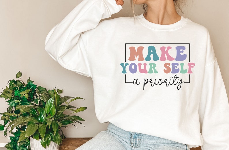 May include: White sweatshirt with the text "MAKE YOUR SELF - a priority" in colorful, retro-style lettering.