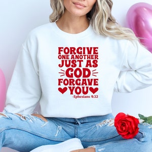 K&ouml;nnte beinhalten: Wei&szlig;er Pullover mit rotem Text, der "Forgive one another just as God forgave you" mit zwei roten Herzen und dem Schriftzitat "Epheser 4:32" lautet.