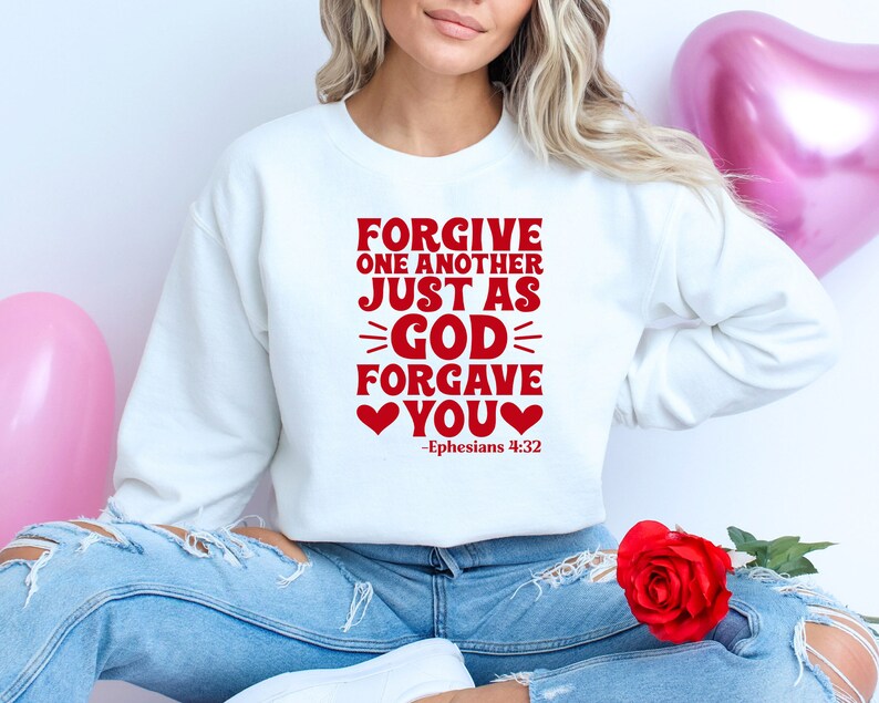 K&ouml;nnte beinhalten: Wei&szlig;er Pullover mit rotem Text, der "Forgive one another just as God forgave you" mit zwei roten Herzen und dem Schriftzitat "Epheser 4:32" lautet.