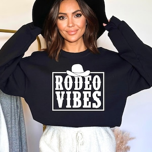 Op de afbeelding: Zwart gecropped sweatshirt met een witte afbeelding van een cowboyhoed en de tekst "Rodeo Vibes".