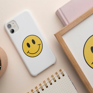 K&ouml;nnte beinhalten: Weisses Handy-Case mit einem gelben Smiley-Grafik. Das Handy-Case hat ein schwarzes Kameraobjektiv.