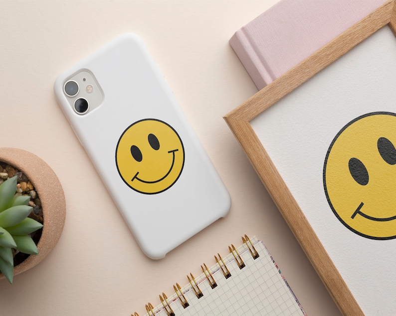 K&ouml;nnte beinhalten: Weisses Handy-Case mit einem gelben Smiley-Grafik. Das Handy-Case hat ein schwarzes Kameraobjektiv.
