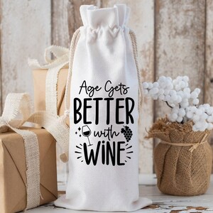 May include: White cotton wine bottle bag with black text that reads "Age Gets Better With Wine" and a small wine glass and grapes graphic.
