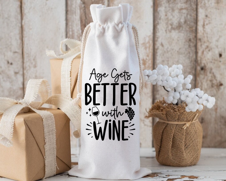 May include: White cotton wine bottle bag with black text that reads "Age Gets Better With Wine" and a small wine glass and grapes graphic.