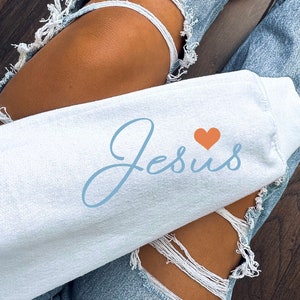 May include: A white sweatshirt with the word "Jesus" written in blue cursive script and a small orange heart above the word.