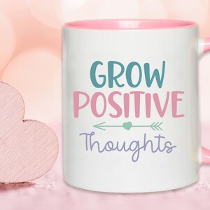 May include: White ceramic coffee mug with a pink rim and handle. The mug has a motivational quote that reads "Grow Positive Thoughts" with an arrow and a heart graphic.