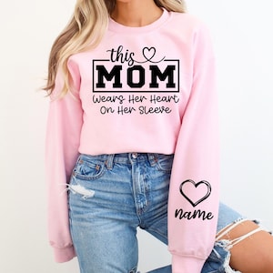 May include: A light pink sweatshirt with the text "this MOM" in a black rectangular box, with a heart above the word "MOM". Below the text is "Wears Her Heart On Her Sleeve". A black heart and the word "name" are on the sleeve.