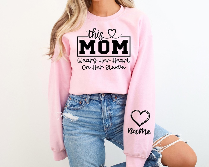 May include: A light pink sweatshirt with the text "this MOM" in a black rectangular box, with a heart above the word "MOM". Below the text is "Wears Her Heart On Her Sleeve". A black heart and the word "name" are on the sleeve.