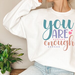 May include: White sweatshirt with the text "you are enough" in colorful lettering with pink hearts.