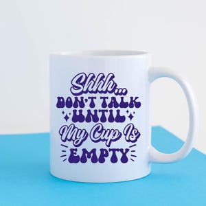 May include: White ceramic mug with a purple design that says "Shhh... Don't talk until my cup is empty".