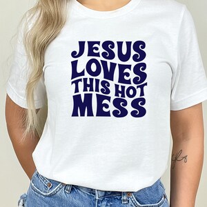 K&ouml;nnte beinhalten: Wei&szlig;es T-Shirt mit einem marineblauen Aufdruck, auf dem "Jesus Loves This Hot Mess" steht.