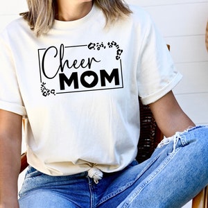 May include: White t-shirt with black text that says "Cheer Mom" in a rectangular frame with a leopard print design.