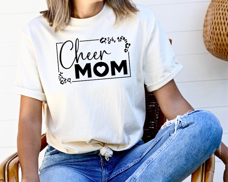 May include: White t-shirt with black text that says "Cheer Mom" in a rectangular frame with a leopard print design.