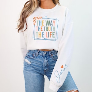May include: White cropped sweatshirt with the text "The Way The Truth The Life" in a colorful frame and a cross. The left sleeve has the word "Jesus" in blue with a small red heart.