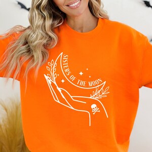 May include: Orange sweatshirt with a white graphic design of a hand holding a crescent moon with stars and leaves. The text "Sisters of the Moon" is written on the moon.