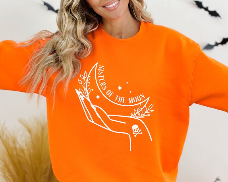 May include: Orange sweatshirt with a white graphic design of a hand holding a crescent moon with stars and leaves. The text "Sisters of the Moon" is written on the moon.