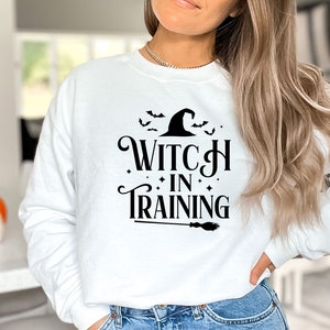 May include: White sweatshirt with black text that reads "Witch in Training" with a witch's hat and bats above the text and a broom below.