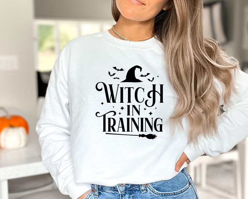 May include: White sweatshirt with black text that reads "Witch in Training" with a witch's hat and bats above the text and a broom below.