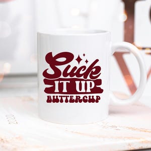 May include: White ceramic coffee mug with a burgundy red design that says "Suck It Up Buttercup".