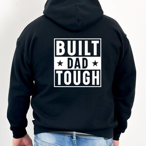 Op de afbeelding: Zwart hoodie met een witte afbeelding op de rug die "BUILT DAD TOUGH" zegt met een ster aan weerszijden van het woord "DAD".
