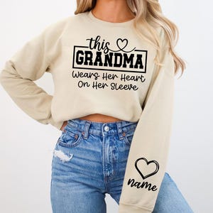 May include: A beige sweatshirt with the text "this GRANDMA Wears Her Heart On Her Sleeve" in black. A black heart graphic is on the sleeve, with space for a name. The sweatshirt is paired with blue jeans.