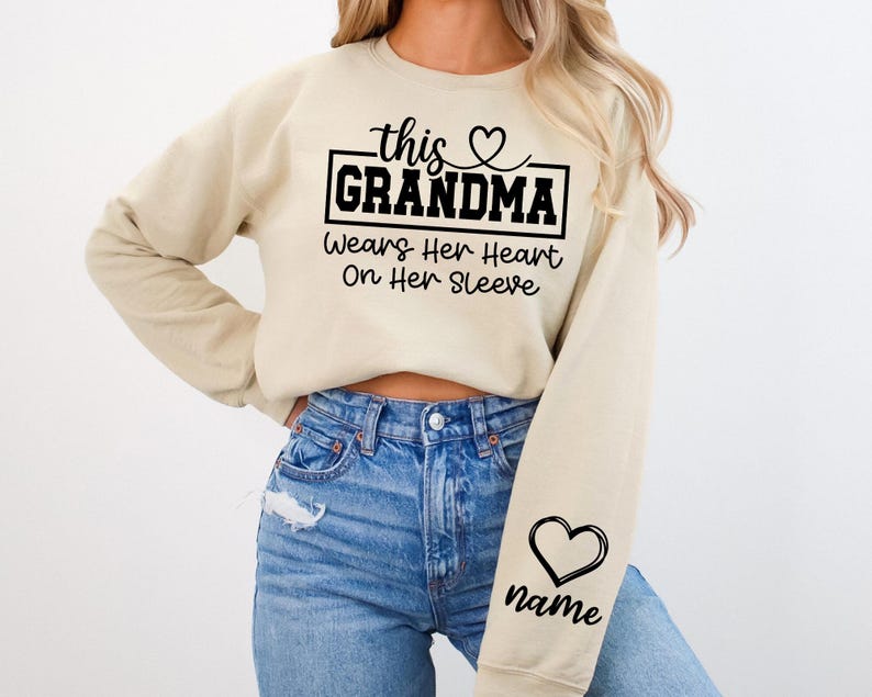 May include: A beige sweatshirt with the text "this GRANDMA Wears Her Heart On Her Sleeve" in black. A black heart graphic is on the sleeve, with space for a name. The sweatshirt is paired with blue jeans.