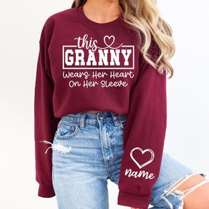 May include: Burgundy crewneck sweatshirt with white text that reads "this GRANNY wears her heart on her sleeve." A white heart and the word "name" are on the sleeve. The sweatshirt is worn with blue jeans.
