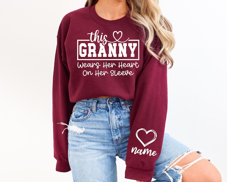 May include: Burgundy crewneck sweatshirt with white text that reads "this GRANNY wears her heart on her sleeve." A white heart and the word "name" are on the sleeve. The sweatshirt is worn with blue jeans.
