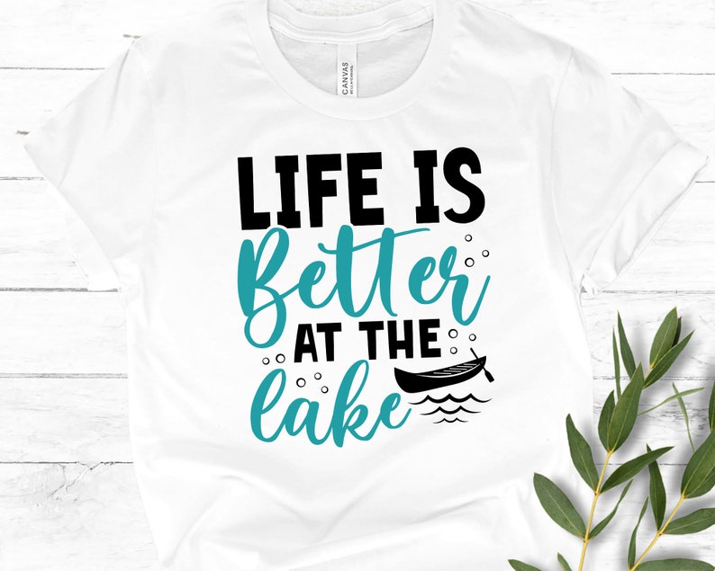 May include: White t-shirt with the text "Life is better at the lake" in black and teal blue lettering. A small black canoe with an oar is pictured next to the text.