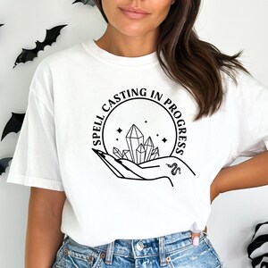 May include: White t-shirt with a black graphic design that says "Spell Casting in Progress" with a hand holding crystals and a snake.