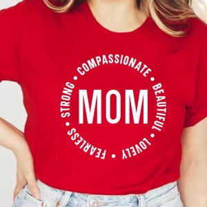 May include: Red t-shirt with a white circle design that says "MOM" in the center and the words "COMPASSIONATE", "BEAUTIFUL", "LOVELY", "FEARLESS", "STRONG" around the circle.