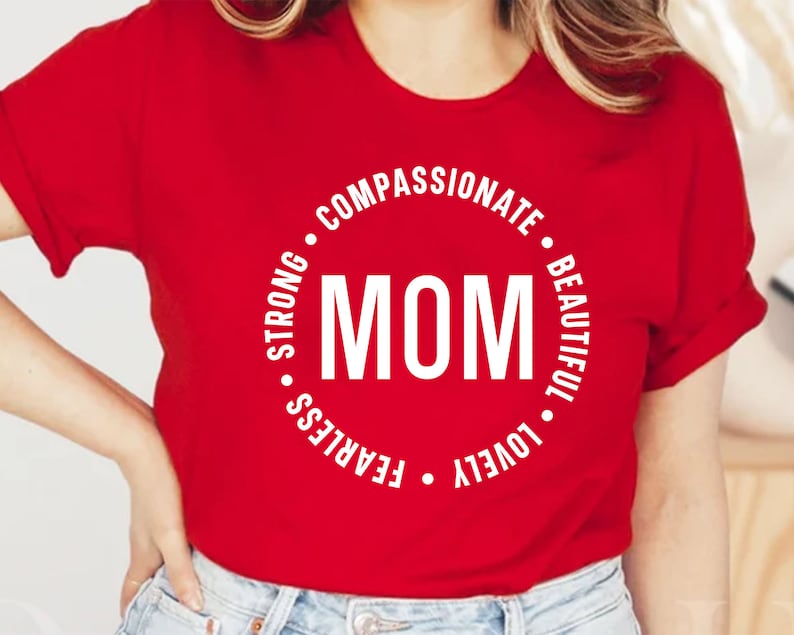 May include: Red t-shirt with a white circle design that says "MOM" in the center and the words "COMPASSIONATE", "BEAUTIFUL", "LOVELY", "FEARLESS", "STRONG" around the circle.