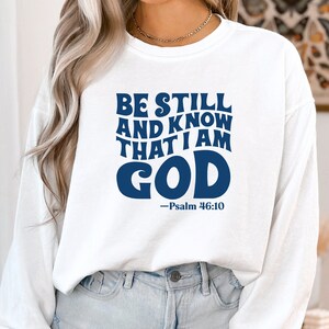 K&ouml;nnte beinhalten: Wei&szlig;er Langarm-Pullover mit einem blauen Aufdruck, der "Be Still and Know That I Am God - Psalm 46:10" lautet.