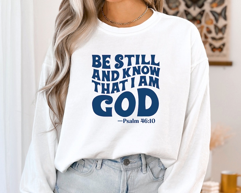 K&ouml;nnte beinhalten: Wei&szlig;er Langarm-Pullover mit einem blauen Aufdruck, der "Be Still and Know That I Am God - Psalm 46:10" lautet.