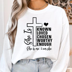 May include: White long-sleeve shirt with a black cross and the text "She Is" and "Known Loved Chosen Worthy Enough She is me i am she".